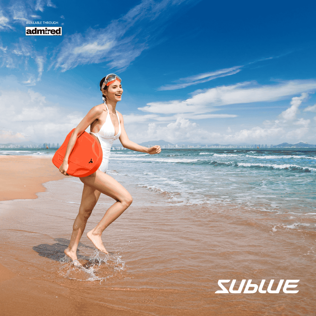 Woman in white swimsuit carrying orange SUBLUE Swii kickboard on the beach.