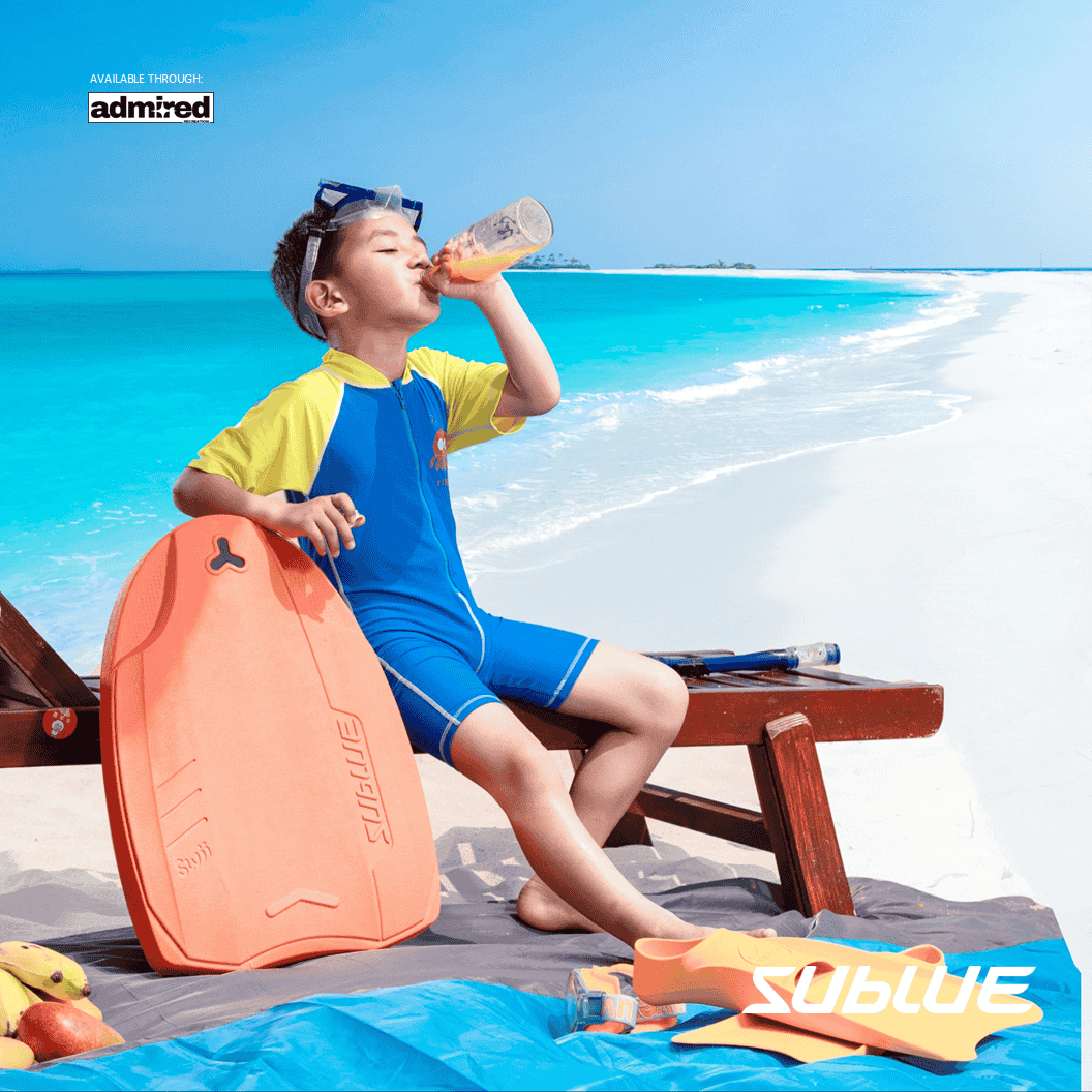 Child in swim gear enjoying a drink with SUBLUE kickboard on a sunny beach.