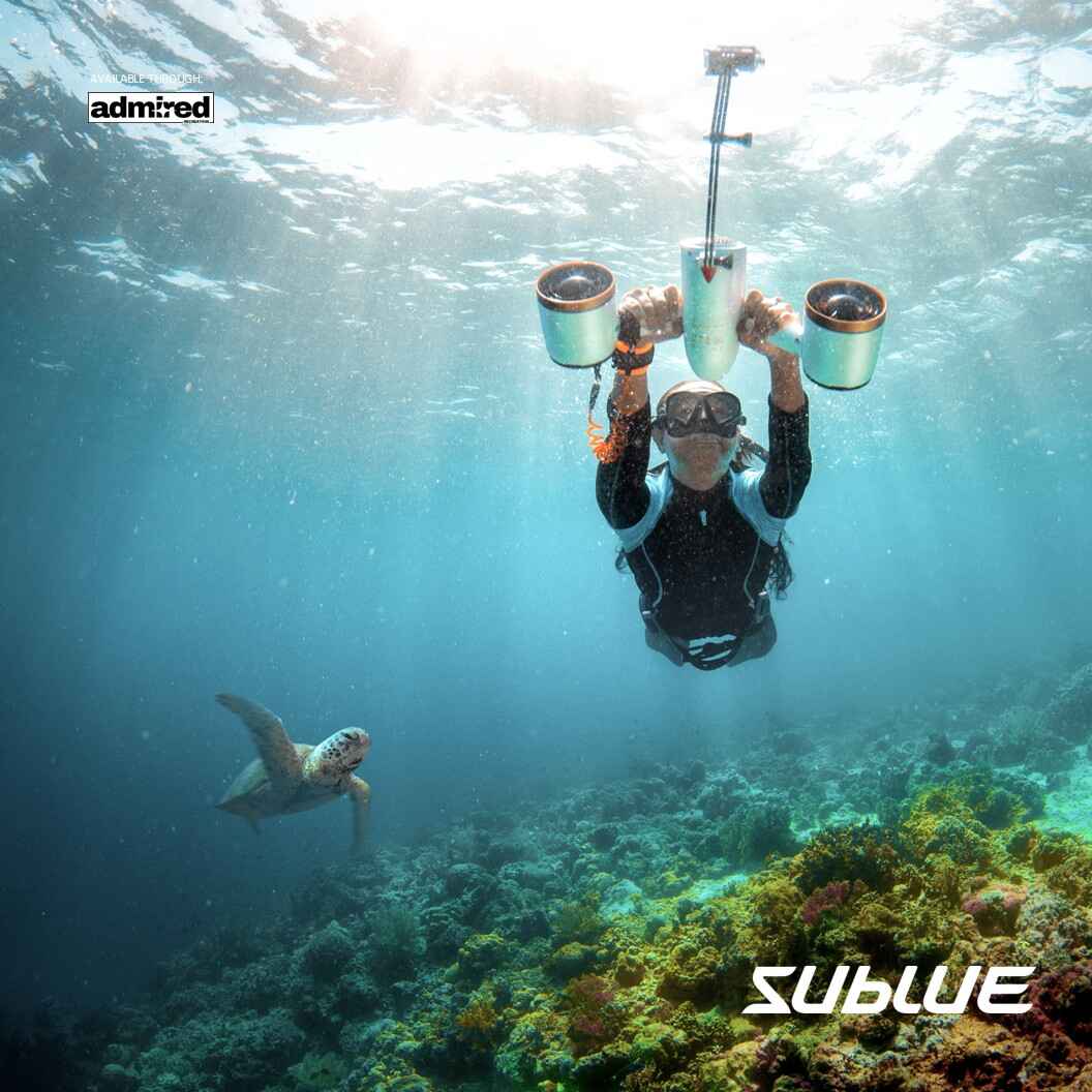 Diver with Sublue scooter and selfie extension stick near coral reef and sea turtle.